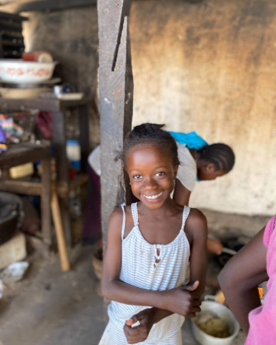 A young African girl is standing in a mud-brick hut with a cheerful smile on her face, seemingly unfazed by her modest surroundings. Her hair is styled with a red headband, and she wears a pink shirt with a cartoon graphic on the front, paired with a patterned skirt. She is standing next to a small bowl containing food, which is being served by an adult, whose hands can be seen pouring something into the bowl behind the girl. Based on the visible context.
